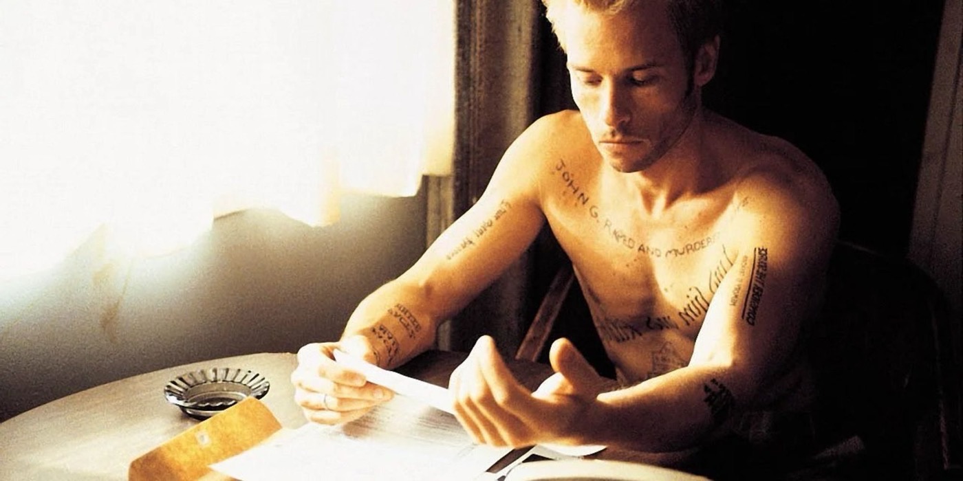 Leonard Shelby sits shirtless at a table reading his notes, his arms and chest covered in tattooed reminders